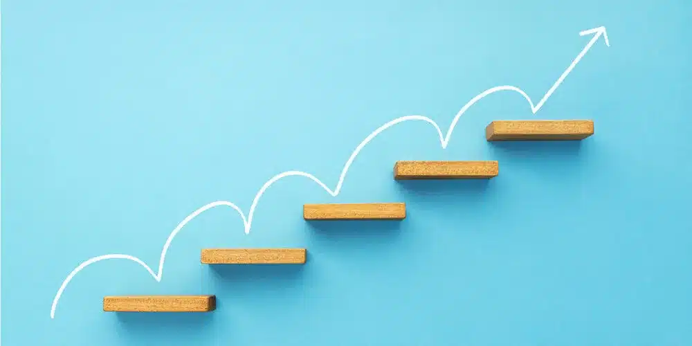 A conceptual image of wooden steps arranged in an upward direction on a blue background, with a white arrow illustrating growth and progress, symbolizing advancement and success.
