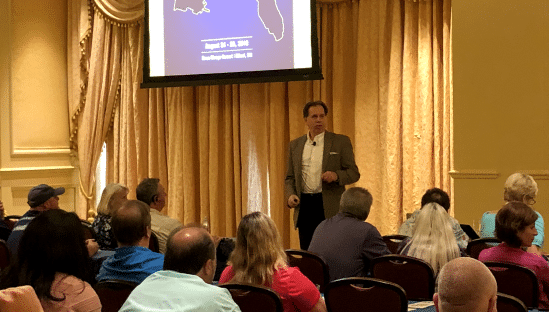 A person is giving an automotive business coaching presentation to a seated audience in a room with beige curtains. A screen displays a map and details.