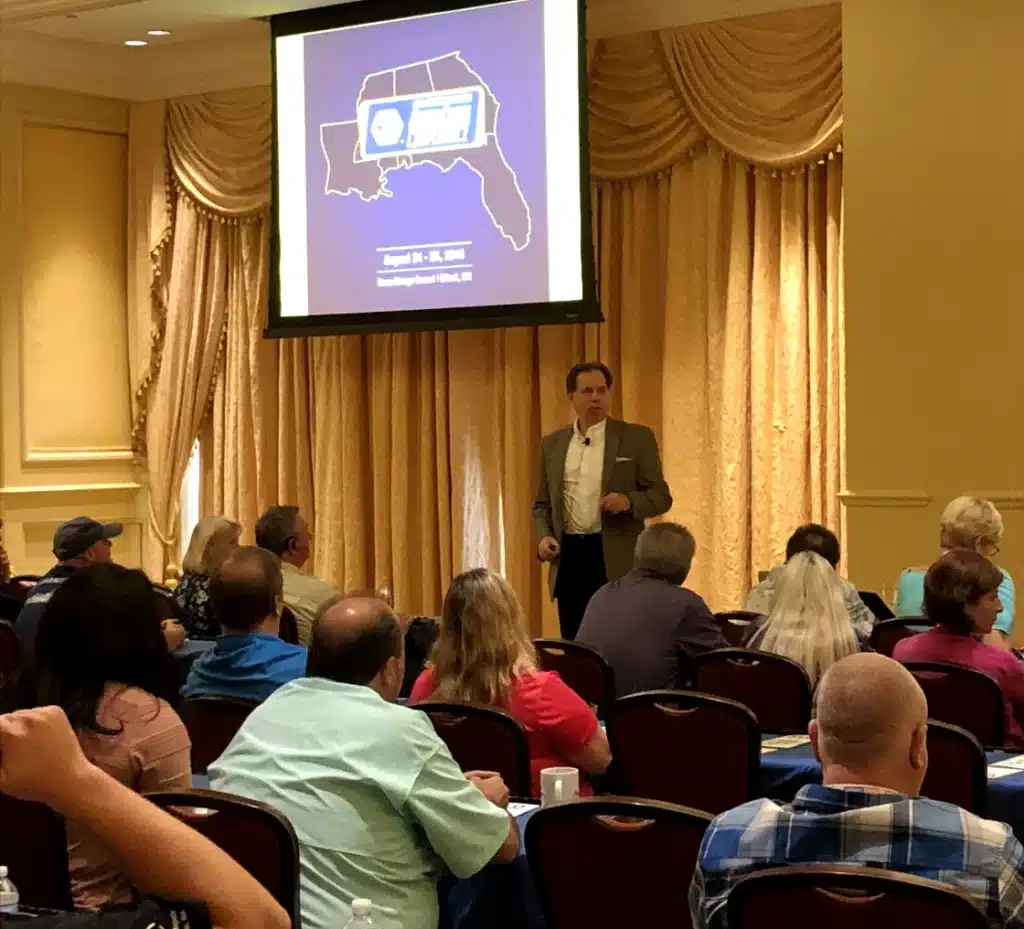 A coach presents a slide on the Southeast US to a seated audience in a conference room with gold curtains.