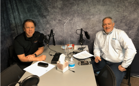 Two men sit at a table with podcast microphones, headphones, and papers in front of them, smiling toward the camera as they discuss coaching strategies for business growth.