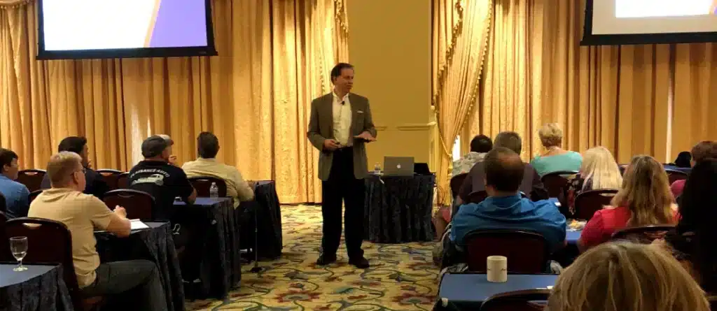 A shop owner in a suit is giving a presentation to an audience in a conference room with golden curtains.