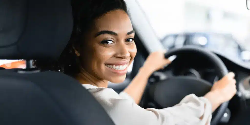 A smiling woman sitting in the driver's seat of a car, looking back towards the camera, symbolizing a positive customer experience in the automotive industry.