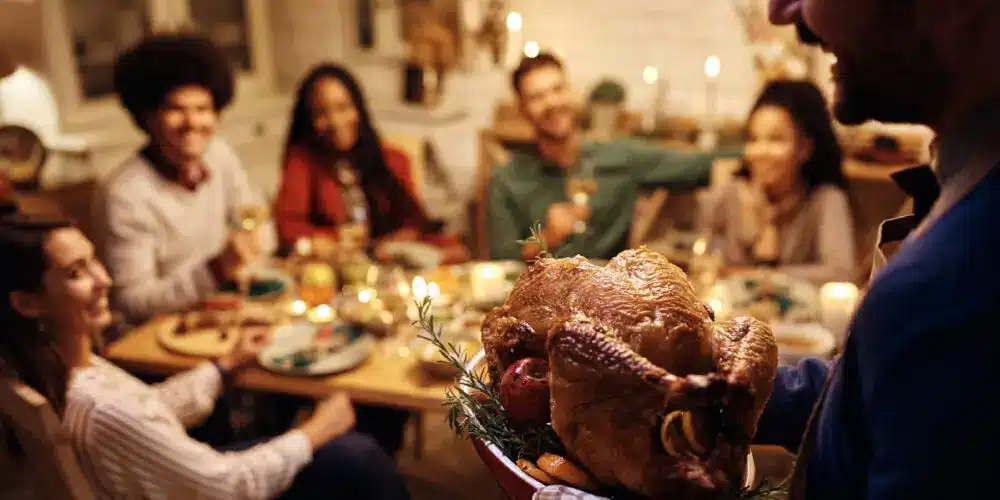 A person carrying a roasted turkey to a festive dinner table, where a group of friends or family members are gathered, smiling and enjoying the celebration in a warmly lit setting.