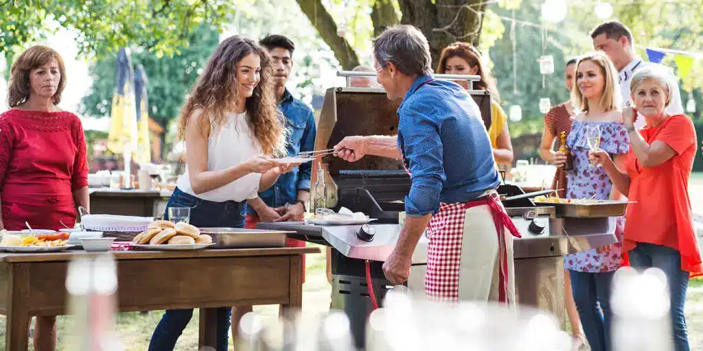 A group of people gathered outdoors at a barbecue, with a man in an apron serving food to a smiling woman, representing community engagement and customer relationship building.