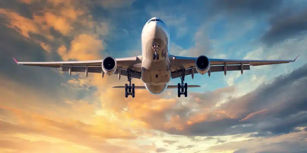 A large commercial airplane flying low with its landing gear extended, set against a dramatic sunset sky with shades of orange, pink, and blue.