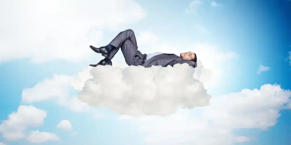 A businessman in a suit is lying comfortably on a fluffy cloud against a bright blue sky, symbolizing relaxation, financial security, or achieving dreams.