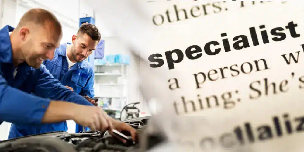 Two mechanics in blue uniforms working on a car engine, with a close-up of the word "specialist" from a dictionary in the background.