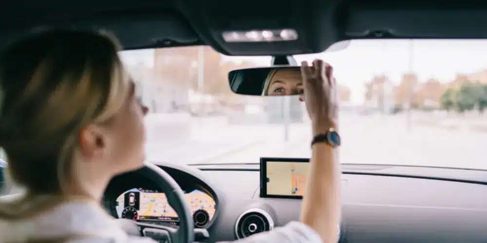 A woman is sitting in the driver's seat of a car, adjusting the rearview mirror while looking into it.